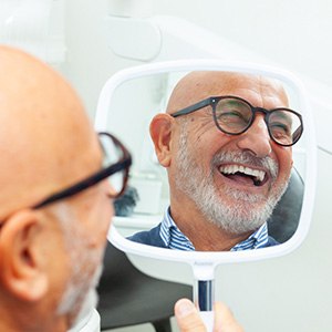 Older bald man in glasses in dental chair smiling at reflection in handheld mirror